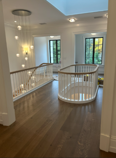 Bright upstairs landing with white railing, hardwood floors, and large windows overlooking trees