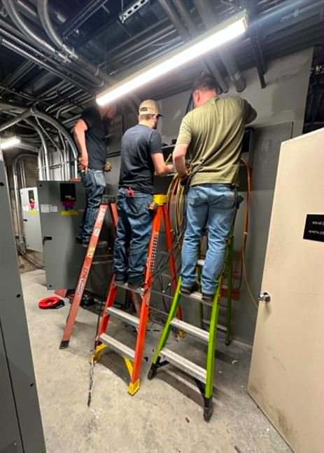 Three workers stand on step ladders fixing overhead pipes in a utility corridor