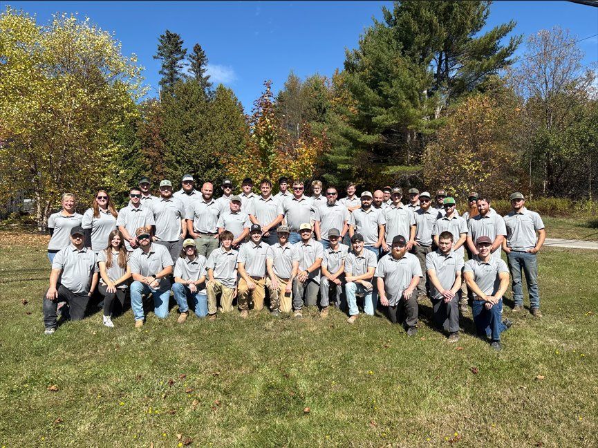 Large group in matching gray shirts posing outdoors on a sunny grassy field with trees in the background