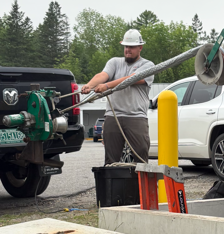 Worker in hard hat operating a green utility machine beside parked trucks and a yellow bollard.