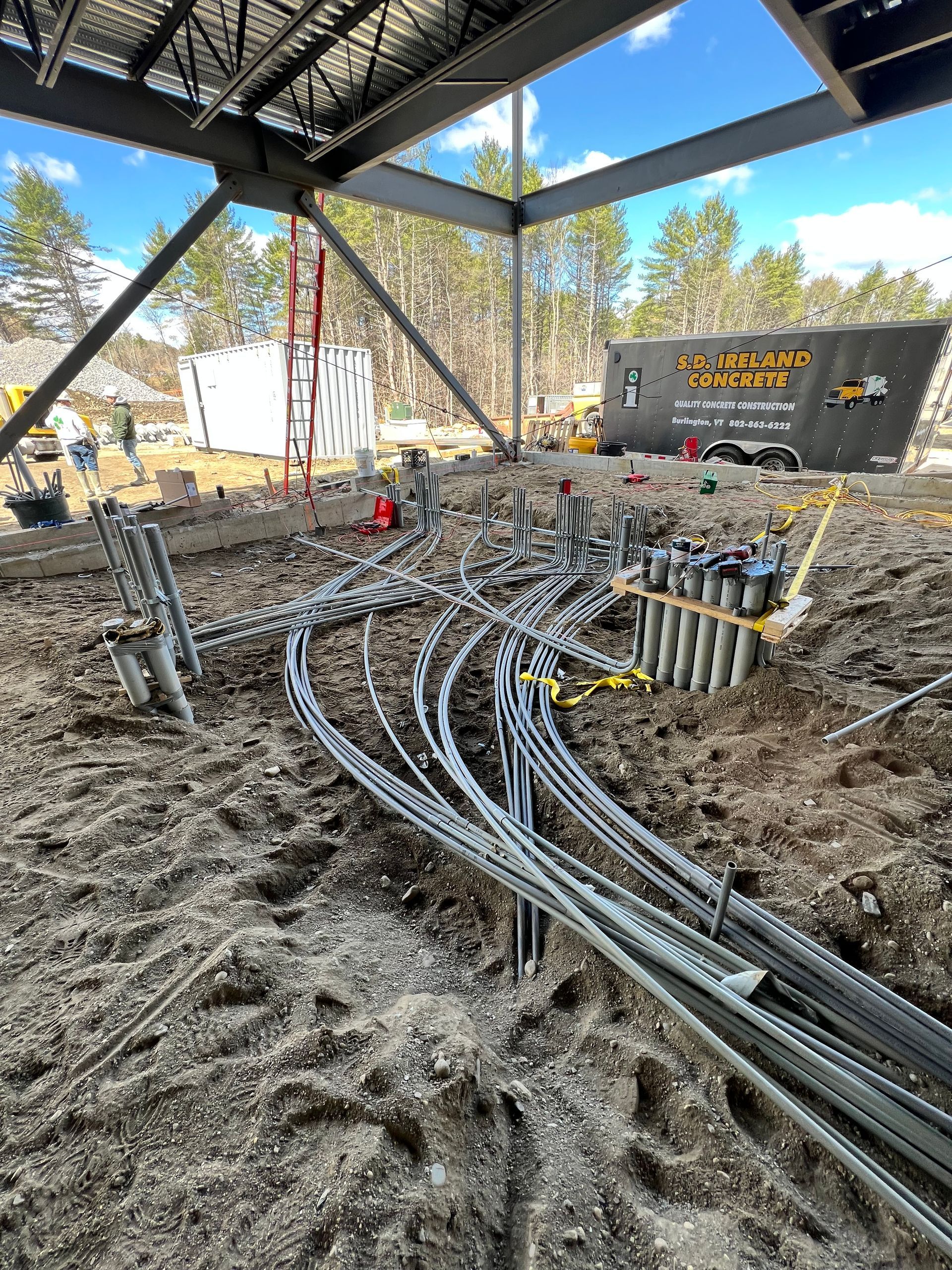 Construction site with tangled cables and conduit under a steel canopy, with machinery and dirt ground.