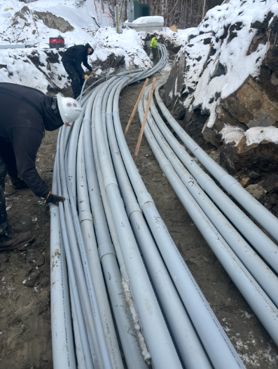Snowy trench with bundled gray pipes laid along a narrow rocky path; workers stand beside them.