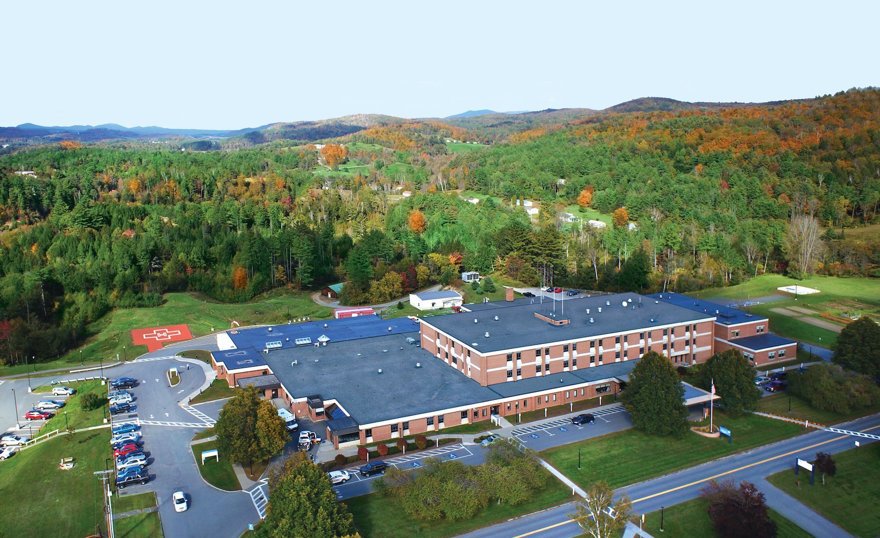 Aerial view of a large brick building with a parking lot, surrounded by green trees and hills in autumn.