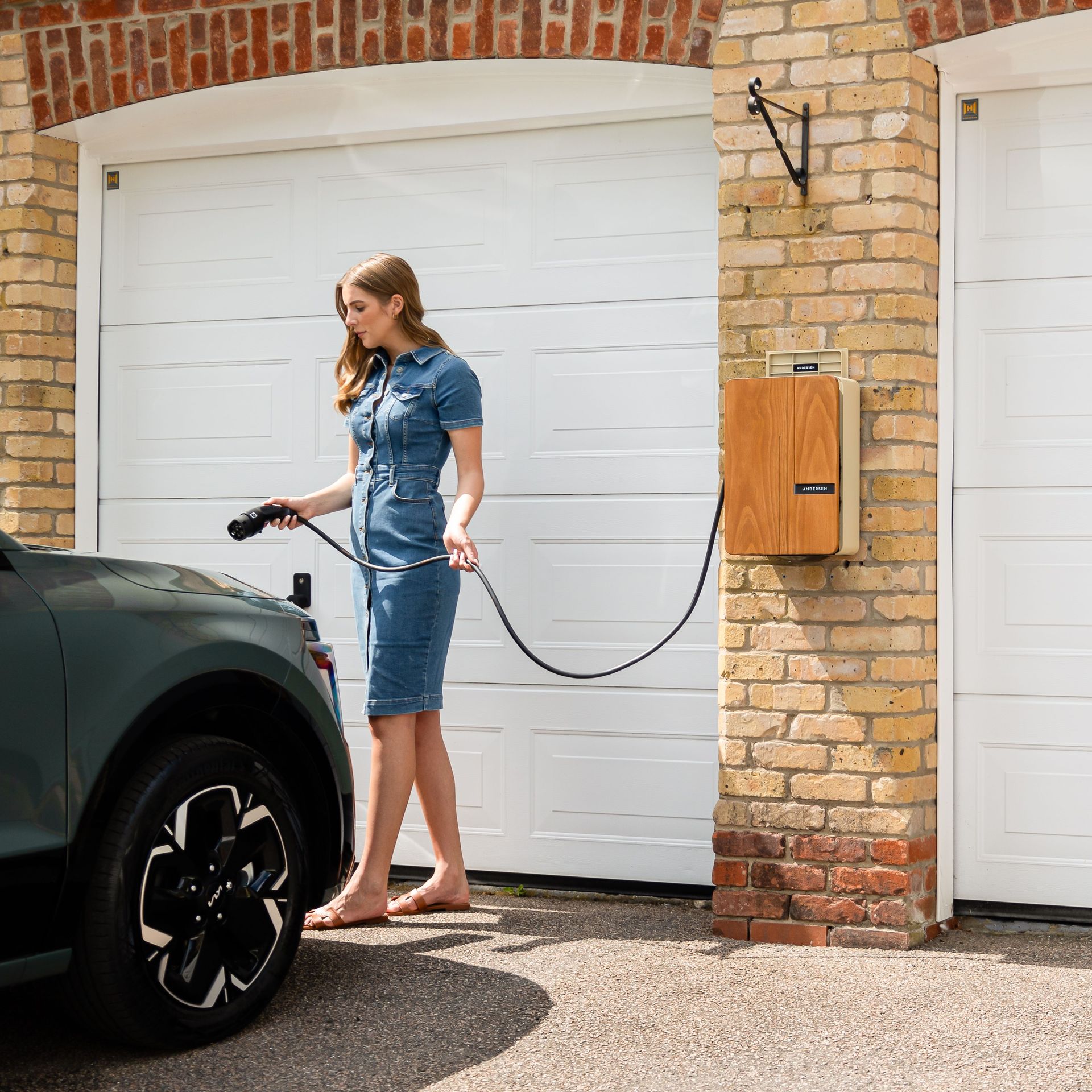 Woman charging an electric SUV in a driveway beside a brick garage door.