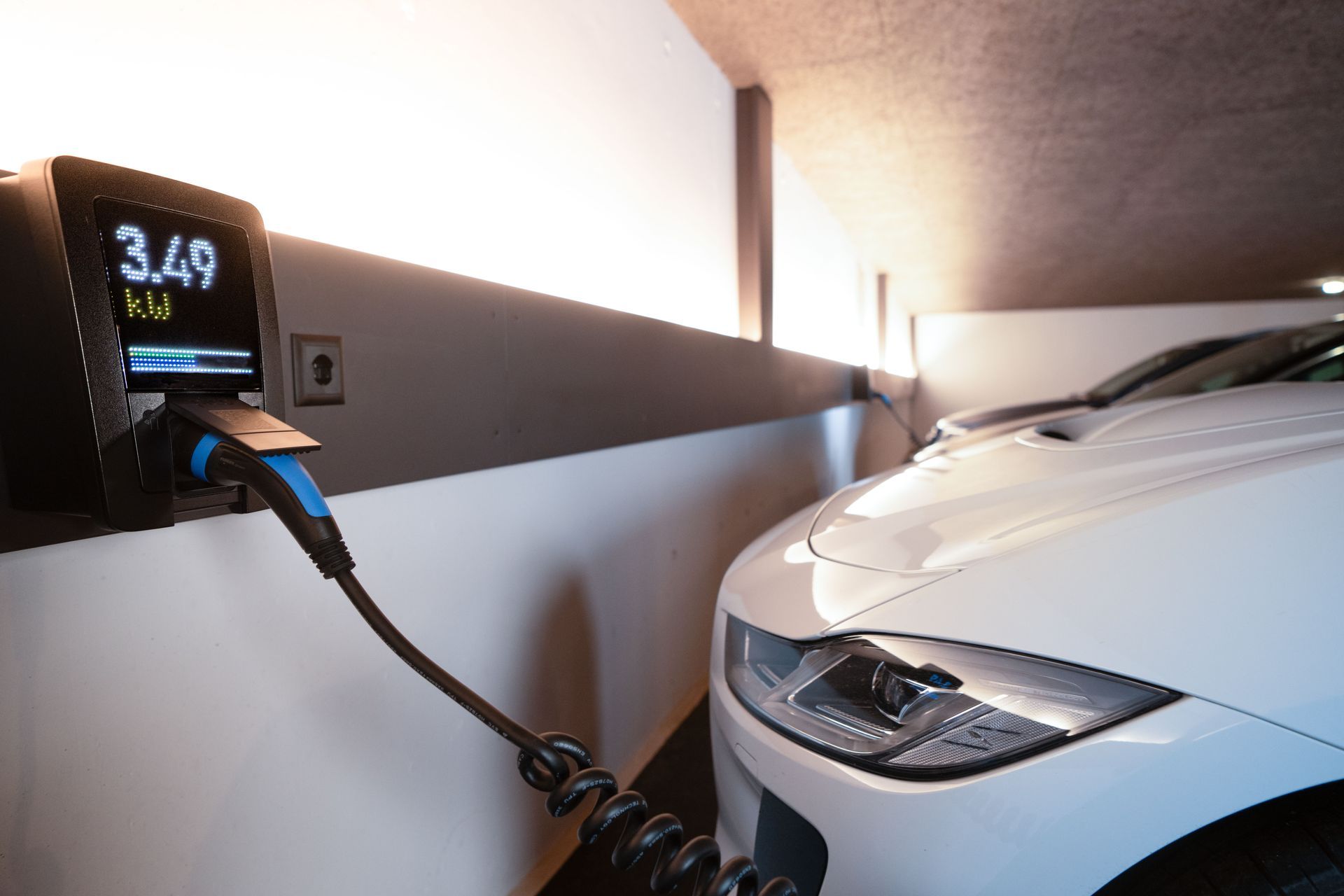 Electric car charging in a garage, with a white vehicle plugged into a wall charger showing 3.49 kW.