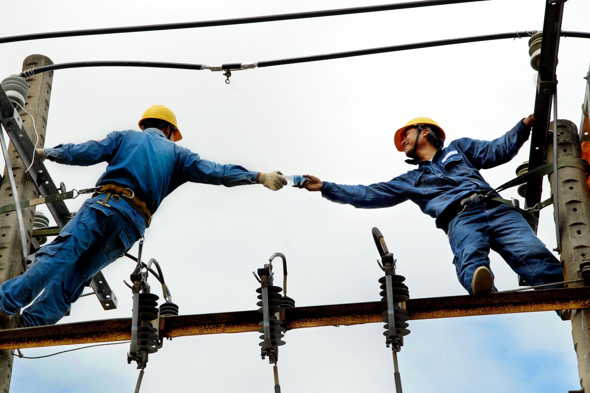 Two workers on a power line reach out to steady each other on a pole high above the ground.