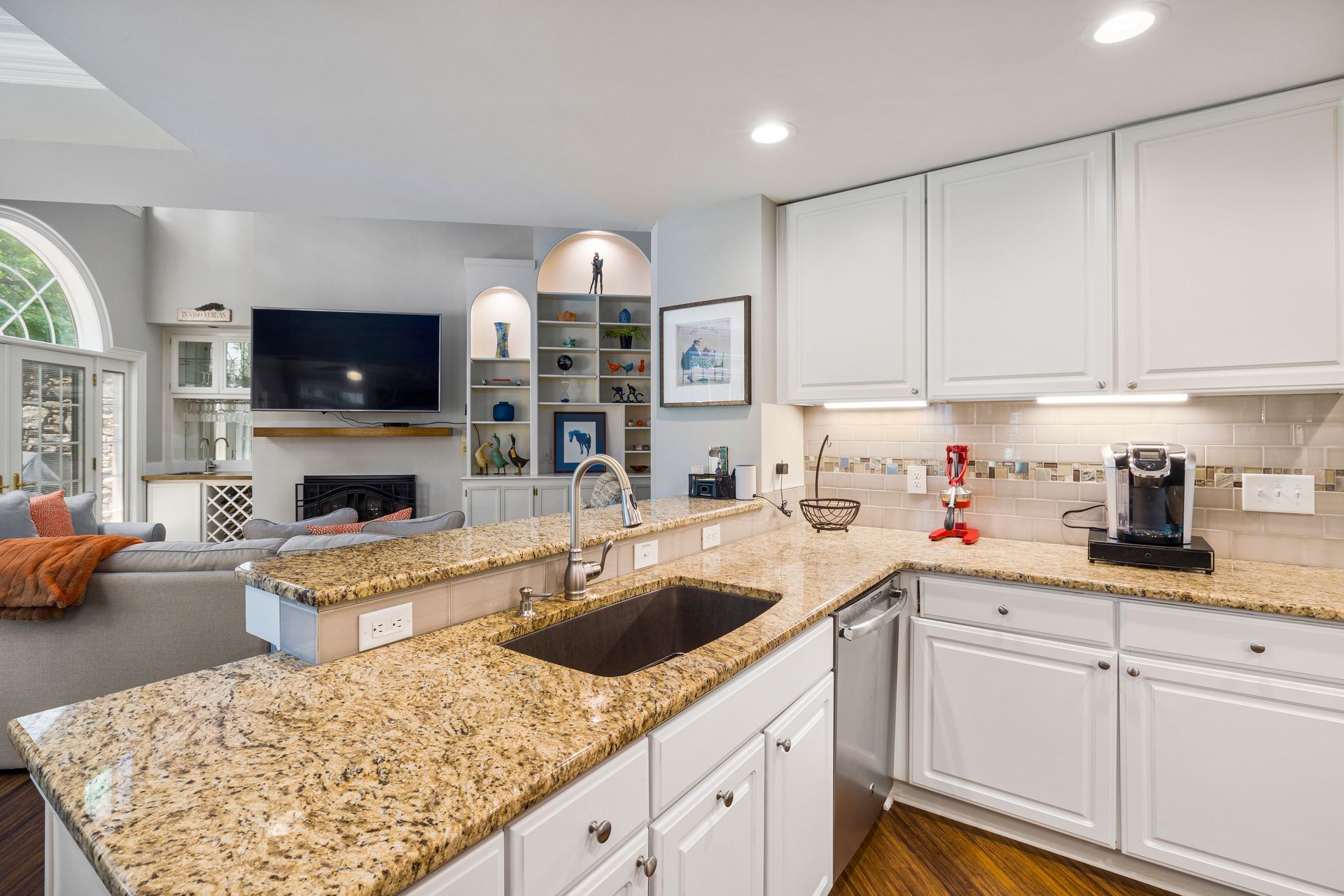 Bright white kitchen with granite countertops, island sink, and built-in shelves beside a living room fireplace.