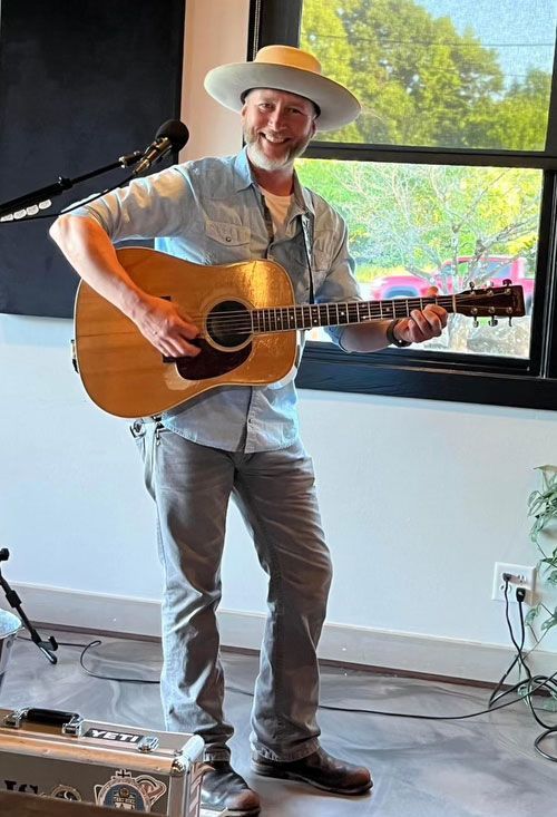 Kris Scott playing an acoustic guitar in front of a window.