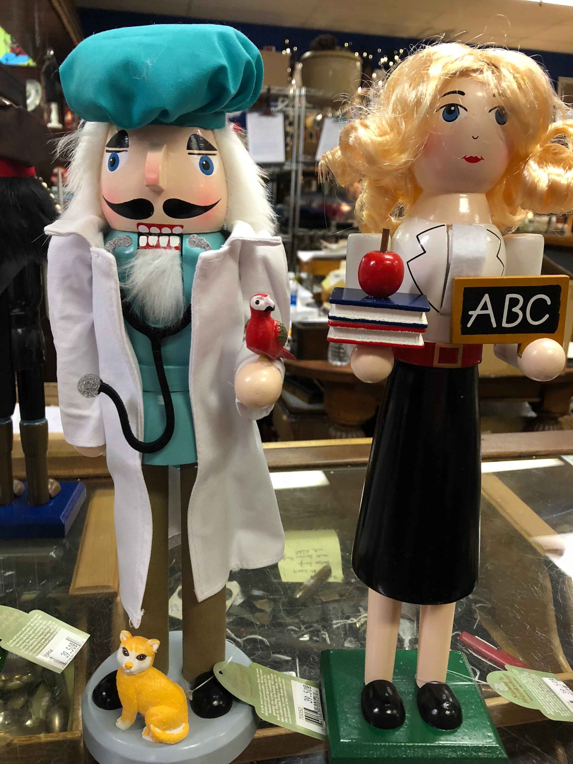 Two handmade figurines in a shop window, one in teal chef attire with a cat, one blonde with a tray and ABC sign.