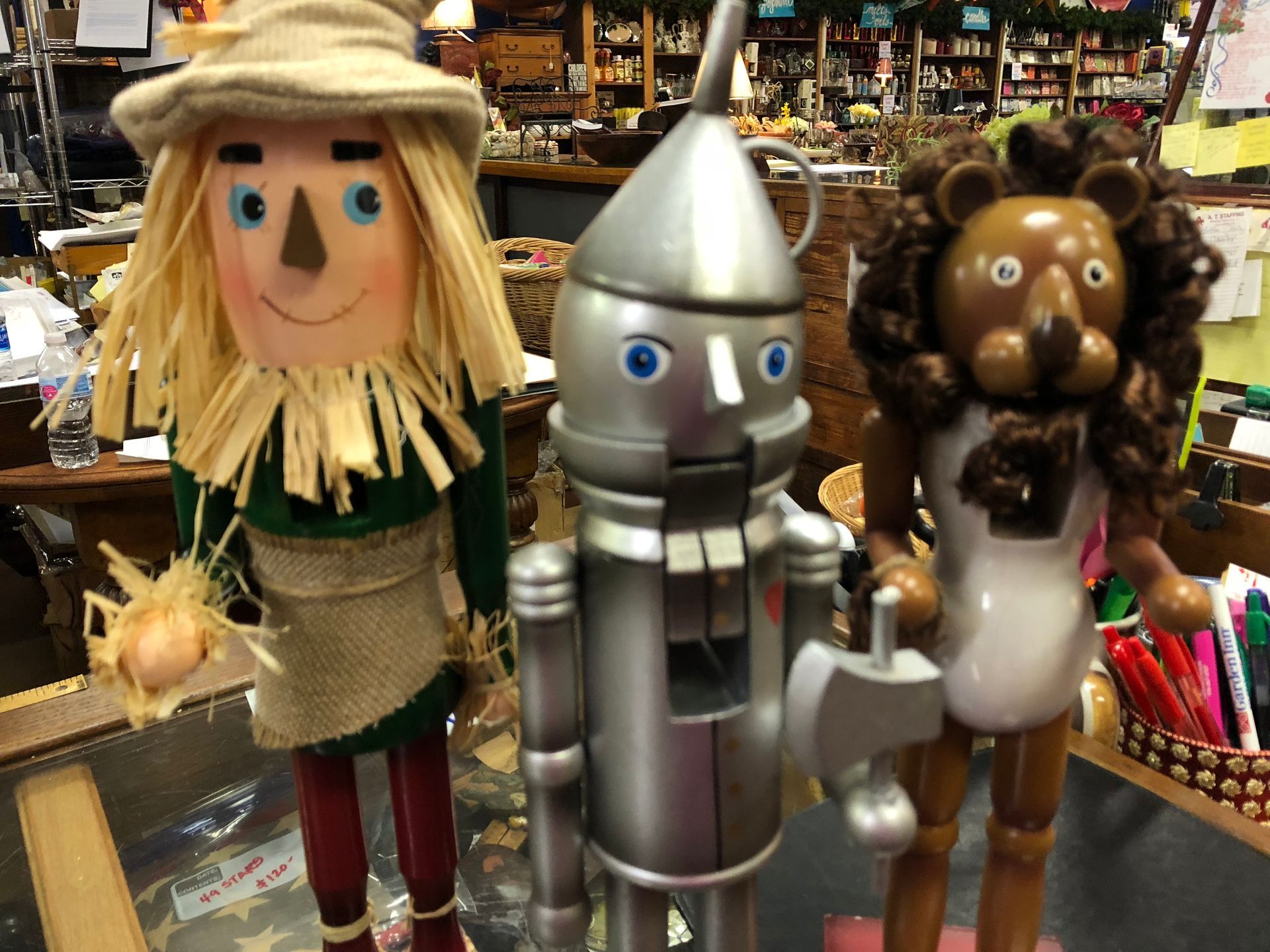 Three whimsical scarecrow dolls on a shop counter, including a silver Tin Man-like figure and a lion-like character.