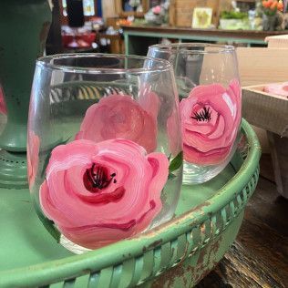 Two clear glasses with pink rose designs sit in a green basket on a shop table.