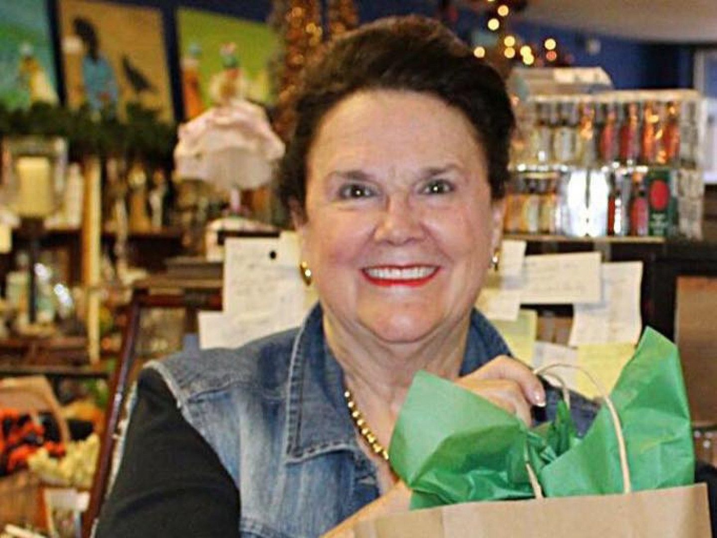 Smiling woman in a denim vest holding a green gift bag in a shop with shelves and lights in the background