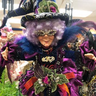 Colorful costumed figure with purple hair, jeweled mask, and feathered hat at a festive event