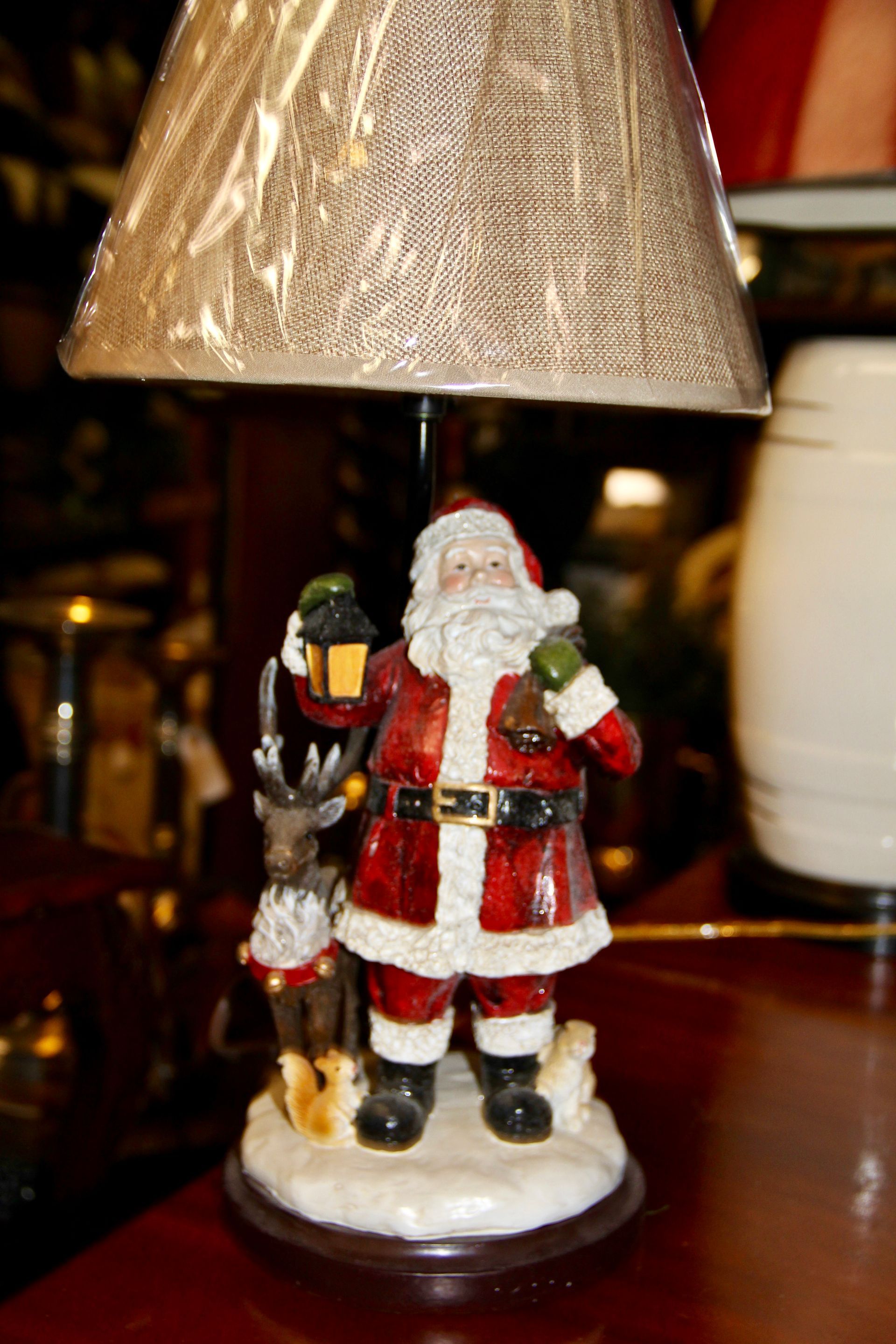 Small Santa figurine holding a lantern, standing on a table beneath a lamp shade.