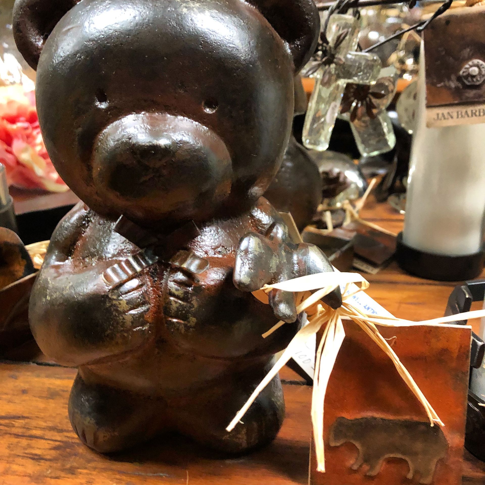 Small dark teddy bear figurine holding a heart, displayed on a wooden table with a wine bottle behind it