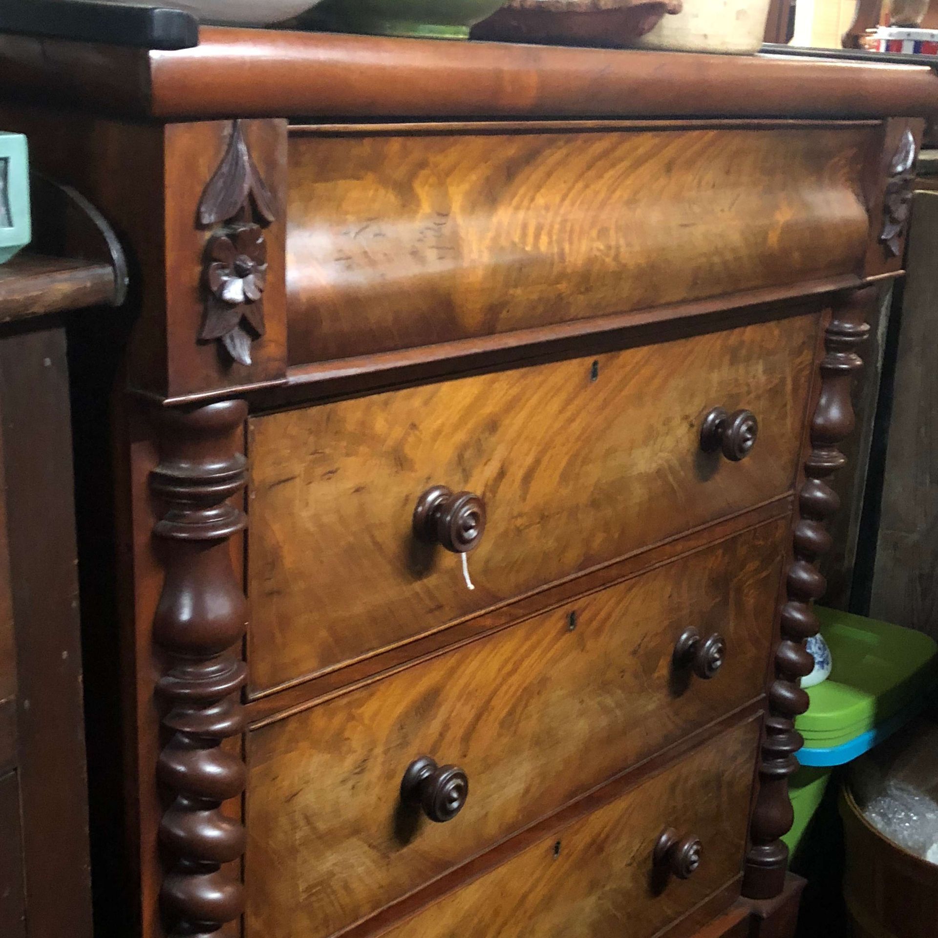 Vintage wooden chest of drawers with carved details and brass knobs in a cluttered room