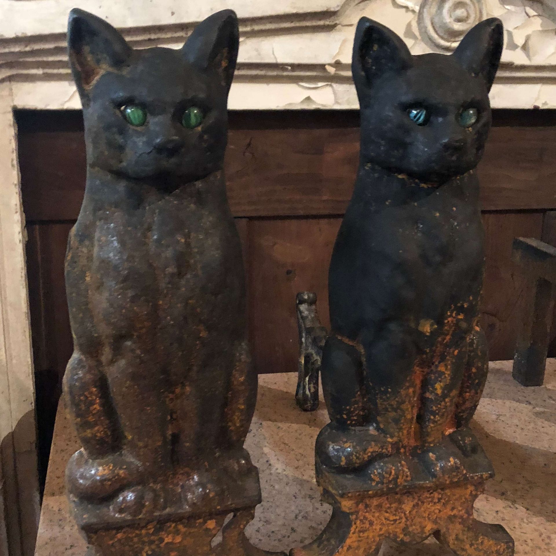 Two black cat statues with green eyes on a mantel, one slightly smaller and rust-stained at the base
