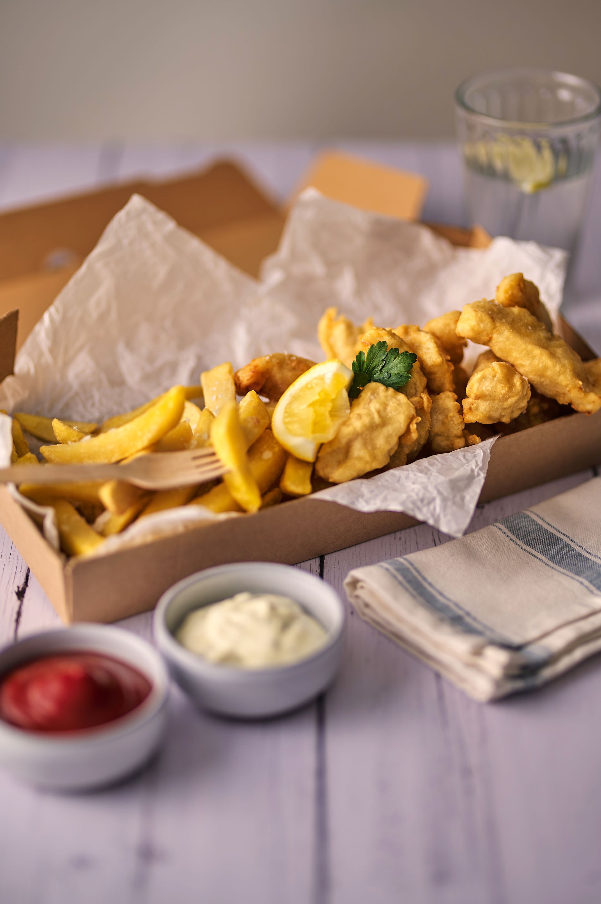 A box of fish and chips with sauces on a table.