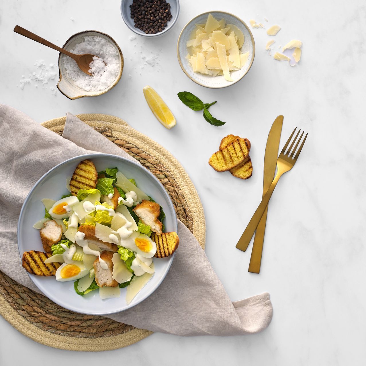 Caesar salad on woven placemat with lemon, cheese, croutons, and gold fork on white table