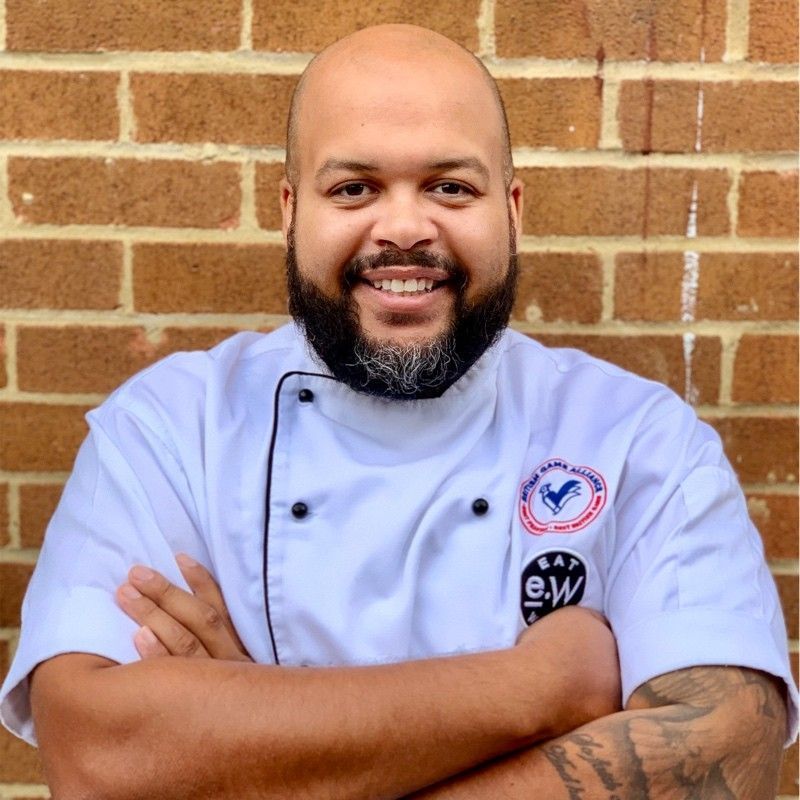 A bald man with a beard wearing a chef 's uniform is standing in front of a brick wall.