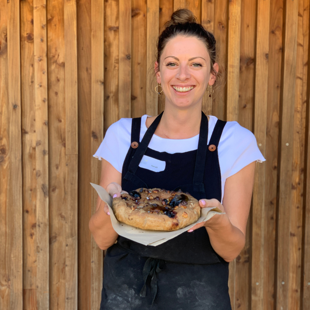 A woman in an apron is holding a pizza in front of a wooden wall.