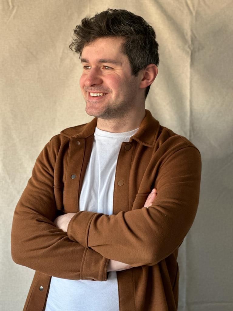 A man in a brown shirt and white shirt is standing with his arms crossed and smiling.