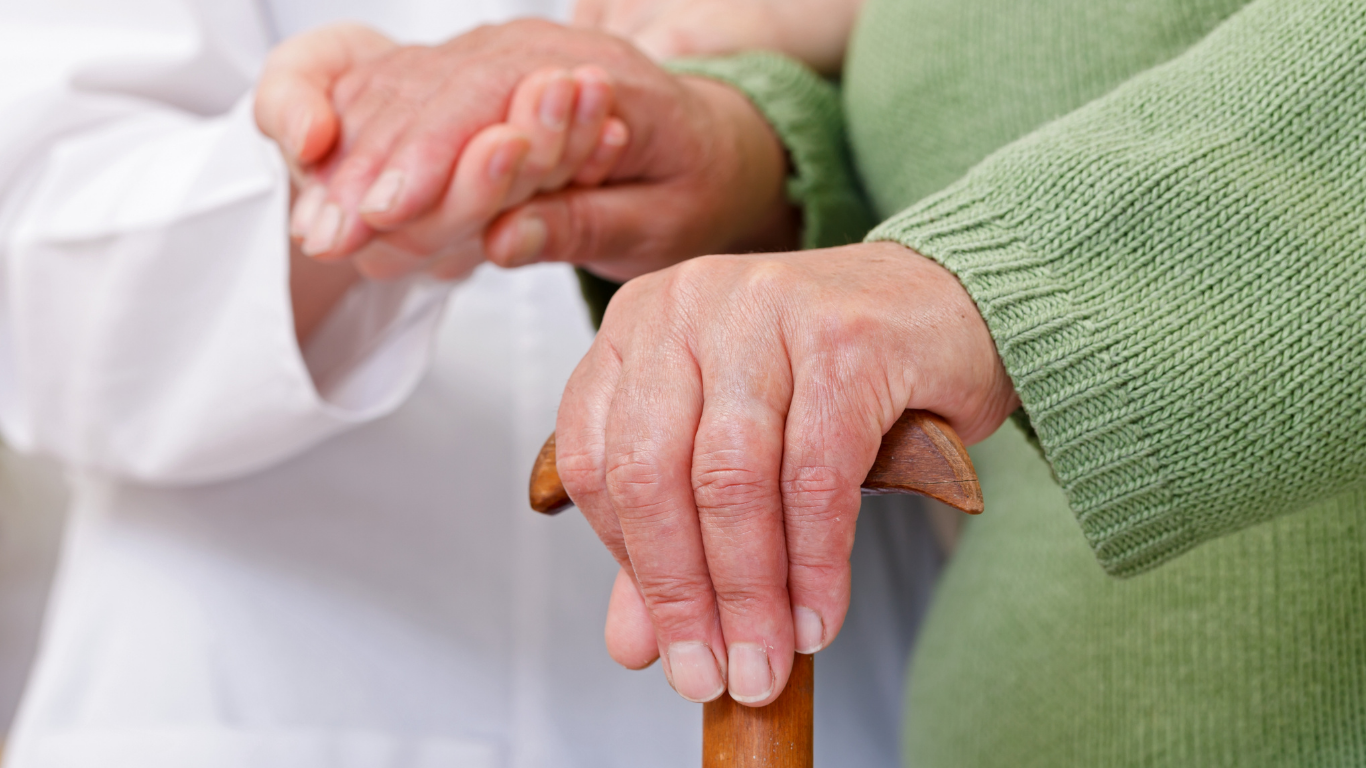 Two hands resting on a wooden cane, one in a green sweater and one in a white sleeve