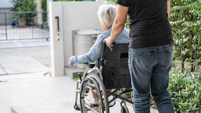 Person pushing someone in a wheelchair along a sidewalk by a building entrance