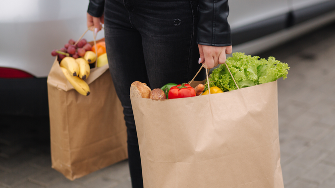 Person carrying paper grocery bags with fresh produce near a car