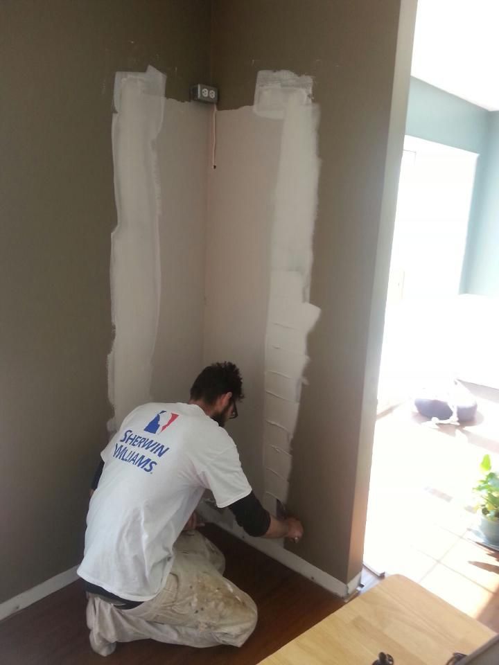 Person, wearing a Sherwin Williams shirt, repairs drywall in a corner of a room.