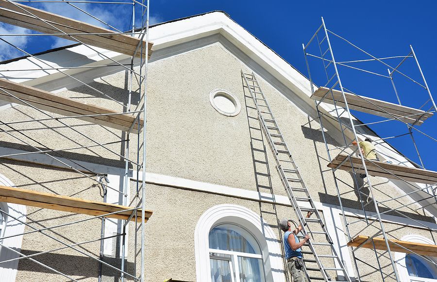 Worker on a ladder beside a stucco building under construction with scaffolding and blue sky