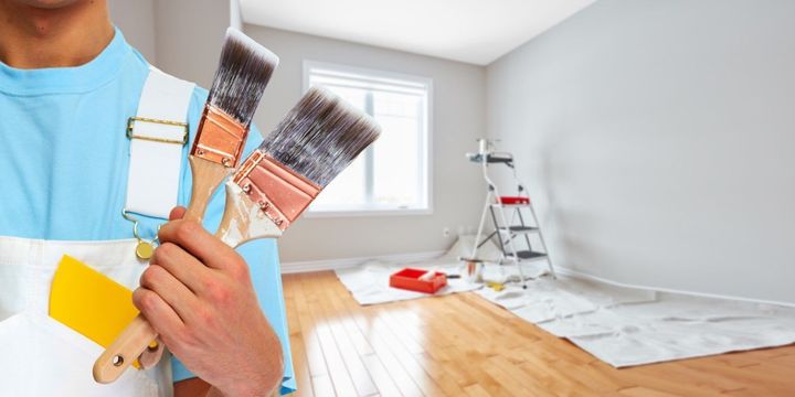 Painter holding brushes and scraper in a bright room with fresh white walls and painting supplies on the floor
