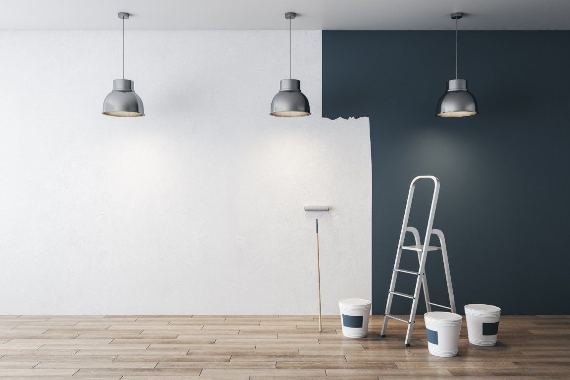 Interior painting in progress: white wall, dark gray accent wall, ladder, paint bucket, and hanging lights.