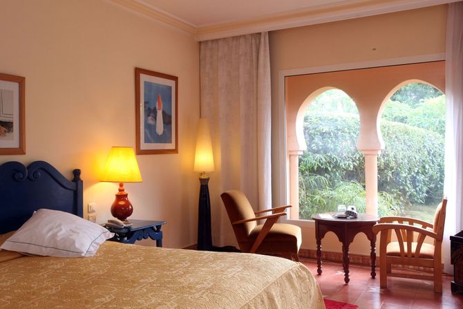 Cozy bedroom with a bed, lamp, two chairs, and arched window view of greenery