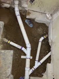 Exposed white PVC plumbing pipes branching in a dirt-lined trench beside a concrete wall