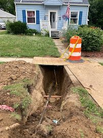 Open trench under a sidewalk by a house, marked with an orange traffic barrel and caution tape.