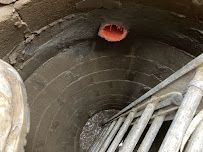 Top view down a narrow concrete well with a red opening at the bottom and metal ladder rails on the right.