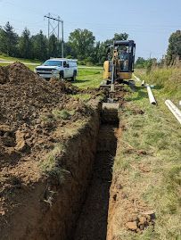 Construction trench beside a grassy road with an excavator, utility truck, and dirt piles