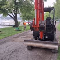 Orange excavator with front blade on a gravel path near a grassy riverbank and trees