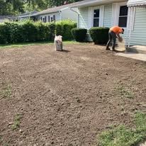 Two workers leveling a bare yard beside a house, with hedges and a white dog nearby