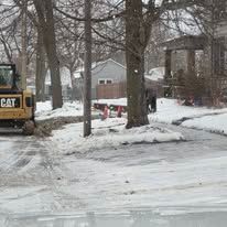 Snowy residential street with a parked CAT machine and bare trees by houses