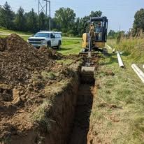 Backhoe digging a trench beside a white pickup truck on a grassy roadside.