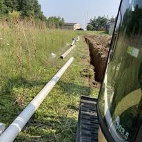 Excavator trenching beside a long white pipe in a grassy field near a house.