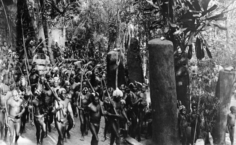 A group of people in traditional attire walk through a wooded area, passing large, dark, upright stone pillars.