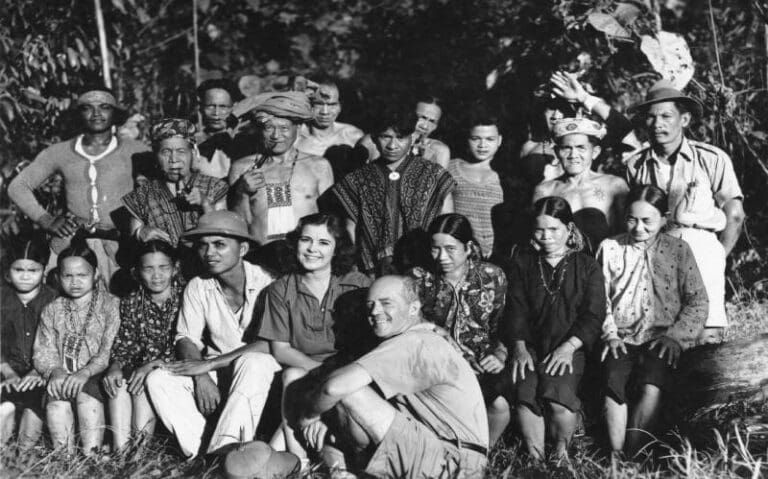 A group of people posed in an outdoor setting, some in traditional attire, with a man crouching in the front center.