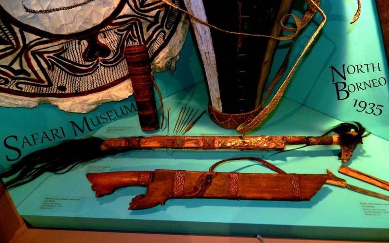 Museum display of North Borneo 1935 artifacts, including a mandau sword, wooden sheath, blowpipe, and woven shield.