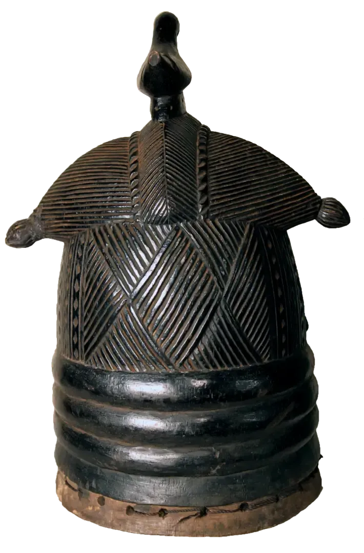 A dark, carved wooden helmet mask with a dome top, geometric incised patterns, and stacked ring details at the base.