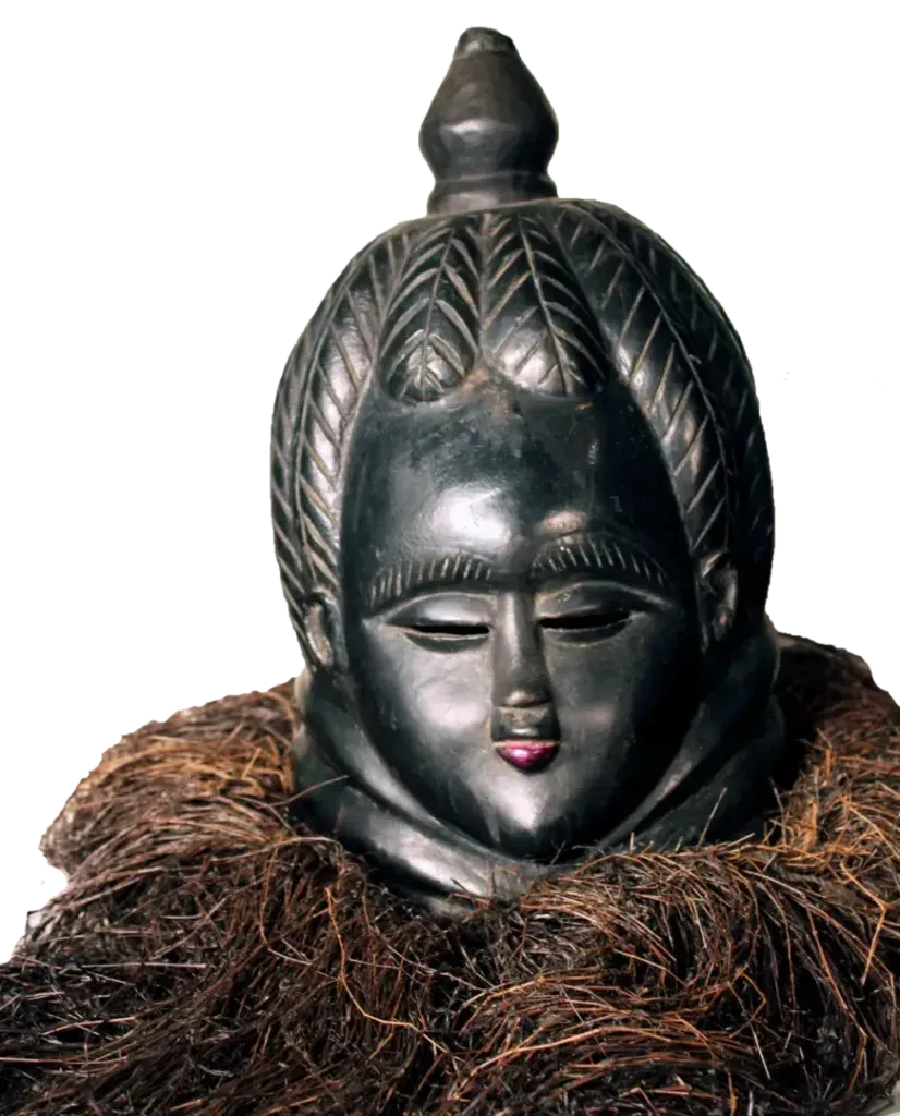 A black, carved wooden helmet mask featuring an ornate hairstyle and a raffia collar against a white background.