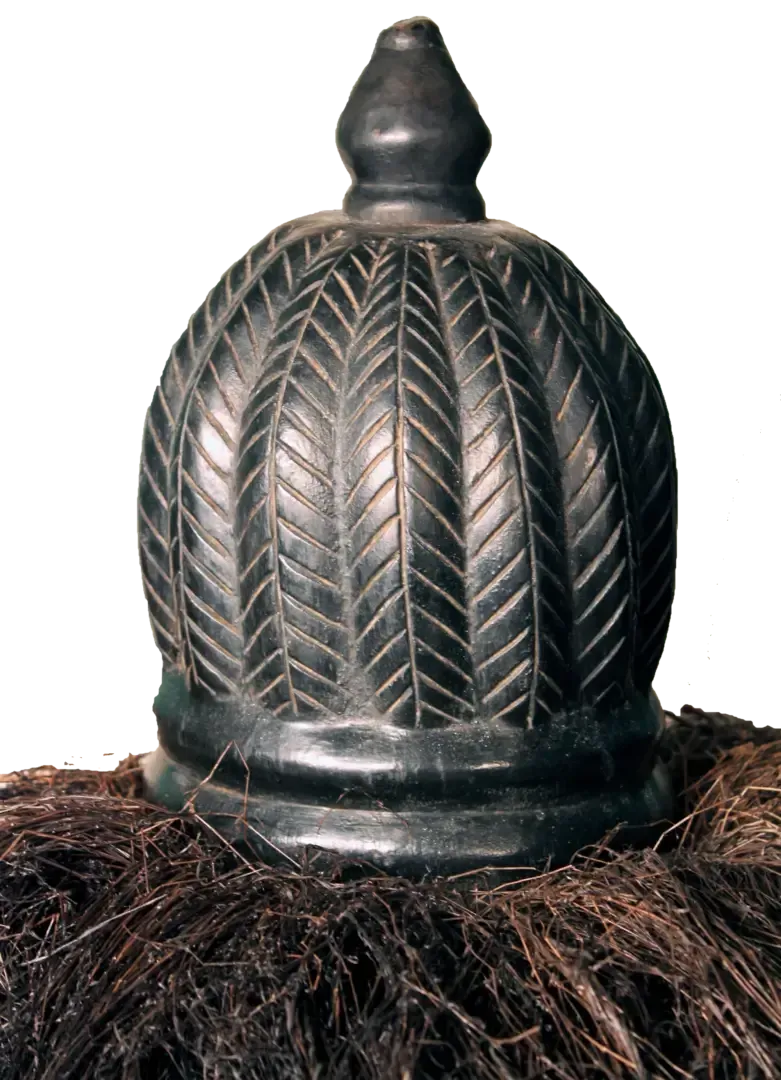A dark, rounded ceremonial headpiece featuring vertical carved herringbone patterns, topped with a small, smooth knob.