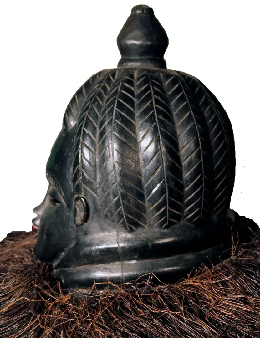 Black carved wooden mask with a chevron-patterned hairstyle and raffia fringe collar against a white background.